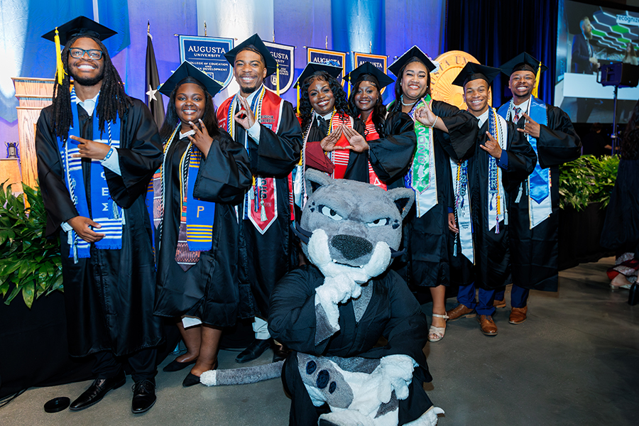 group of male and female students in graduation robes doing hand signs with a cat mascot sitting in front of them 