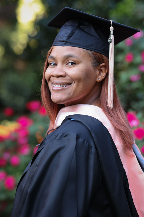 Young woman clothed in graduate regalia and cap standing in front of flower bushes.