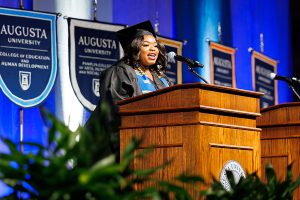 female student in graduation robe speaking at a podium