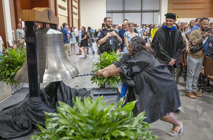 a female student in a graduation robe rings a big silver bell 