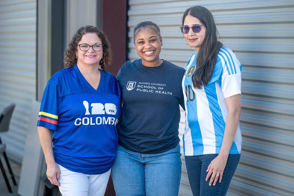 Two women wearing soccer jerseys and one woman wearing a t-shirt that reads, "Augusta University School of Public Health" pose and smile at the camera.