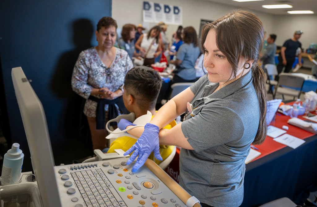 A woman wearing gloves checks something on a screen while performing a carotid artery check on a man.