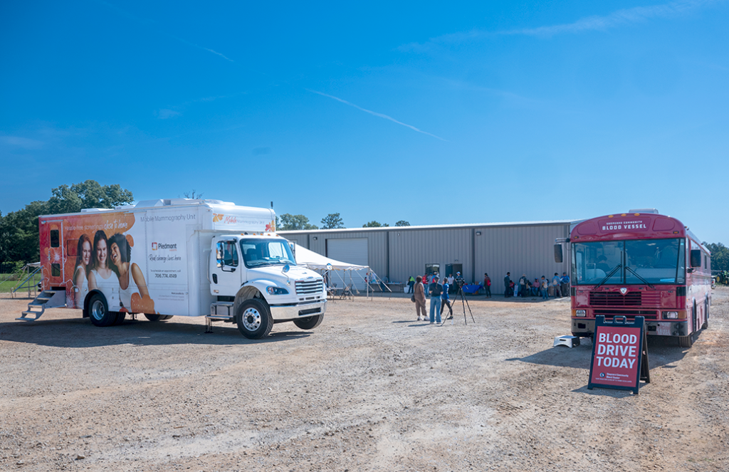 A blood donation mobile and a mobile mammography unit sit in a gravel lot of a health fair.