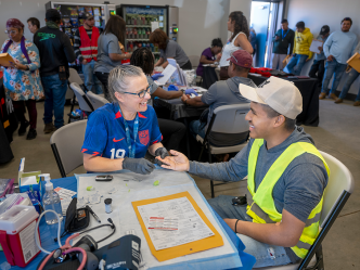A man and woman set a table at a rural health fair. The woman is going to check the man's hemoglobin and they're smiling at each other.
