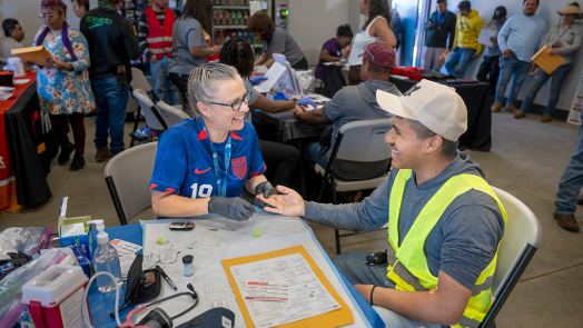 A man and woman set a table at a rural health fair. The woman is going to check the man's hemoglobin and they're smiling at each other.