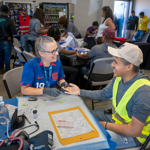 A man and woman set a table at a rural health fair. The woman is going to check the man's hemoglobin and they're smiling at each other.