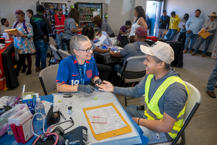 A man and woman set a table at a rural health fair. The woman is going to check the man's hemoglobin and they're smiling at each other.