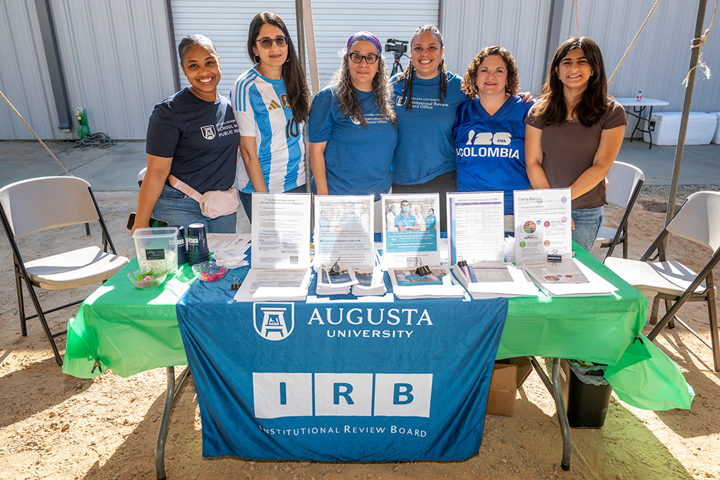 A group of six women stand behind a table at a health fair and smile.