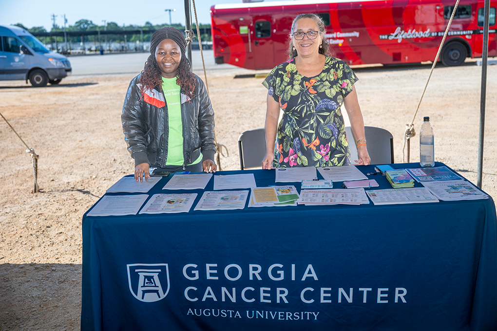 Two women stand behind a Georgia Cancer Center table at a health fair and smile.
