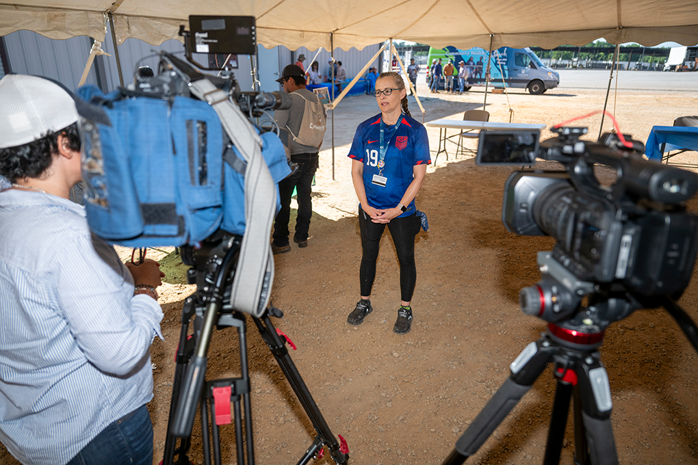 A woman wearing a soccer jersey is interviewed by news crews at a health fair.