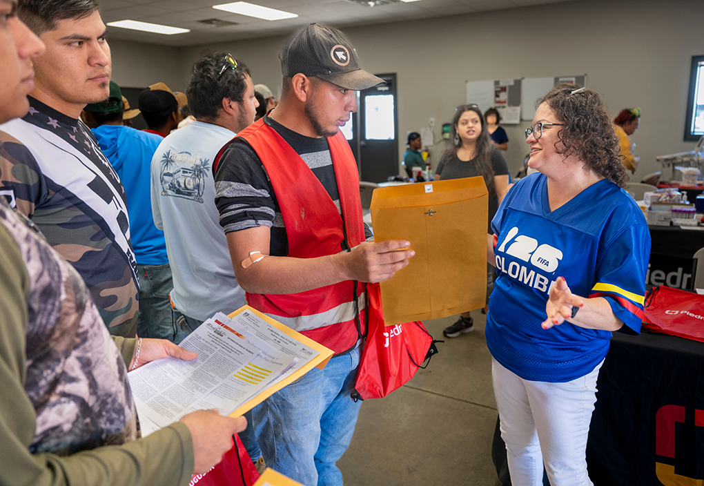 A woman wearing a soccer jersey speaks to men in line at a health fair.