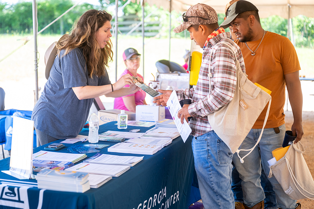 A woman standing behind a table at a health fair talks to two men and hands them a sheet of paper.