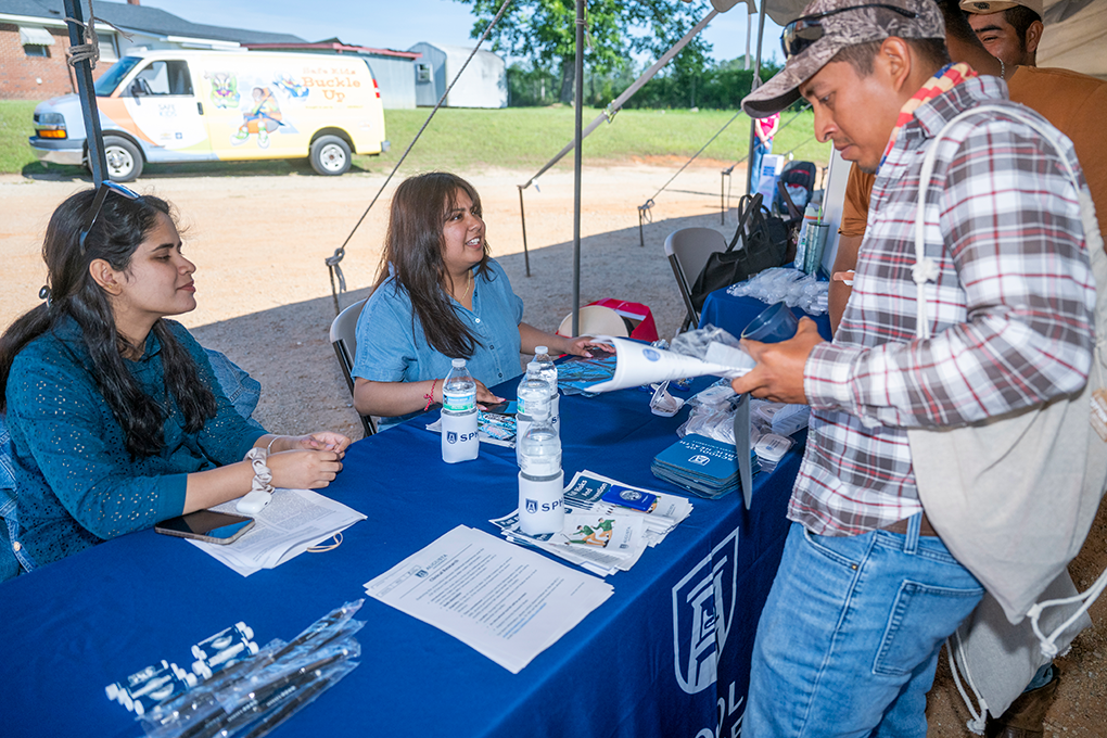 A woman sitting behind a table at a health fair talks to a man and hands him a sheet of paper.