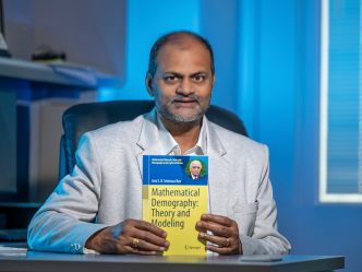 A man sits at a desk and holds up a textbook that reads, "Mathematical Demography: Theory and Modeling."