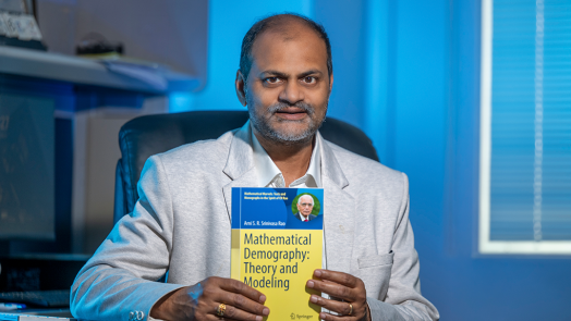 A man sits at a desk and holds up a textbook that reads, "Mathematical Demography: Theory and Modeling."