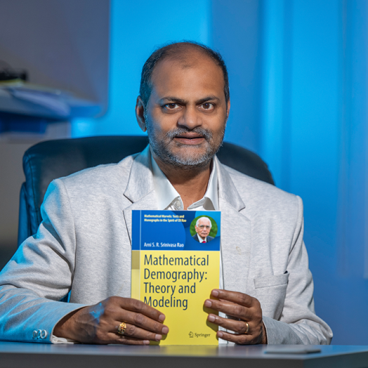 A man sits at a desk and holds up a textbook that reads, "Mathematical Demography: Theory and Modeling."