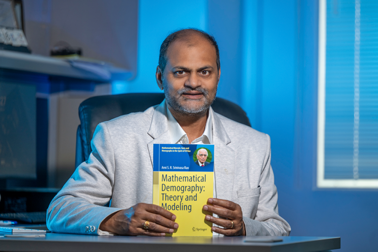 A man sits at a desk and holds up a textbook that reads, "Mathematical Demography: Theory and Modeling."