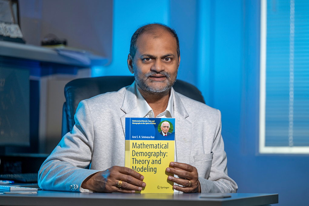 A man sits at a desk and holds up a textbook that reads, "Mathematical Demography: Theory and Modeling."