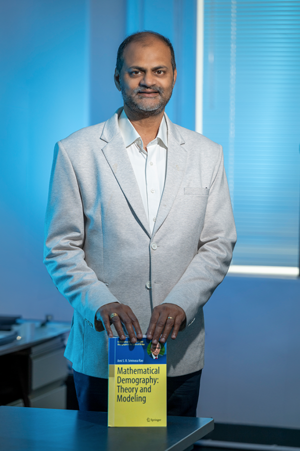 A man wearing a suit stands behind a desk and holds a textbook about mathematical demography in front of him.
