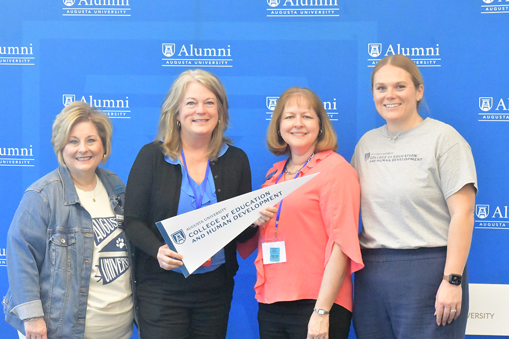 Four women posing for a photo in front of a backdrop with one holding a banner