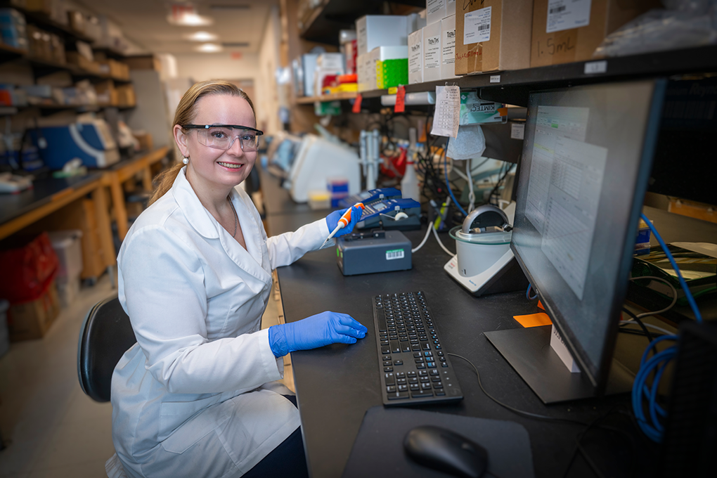 A female scientist wearing a lab coat, gloves and safety glasses sits in front of a computer and smiles at the camera.