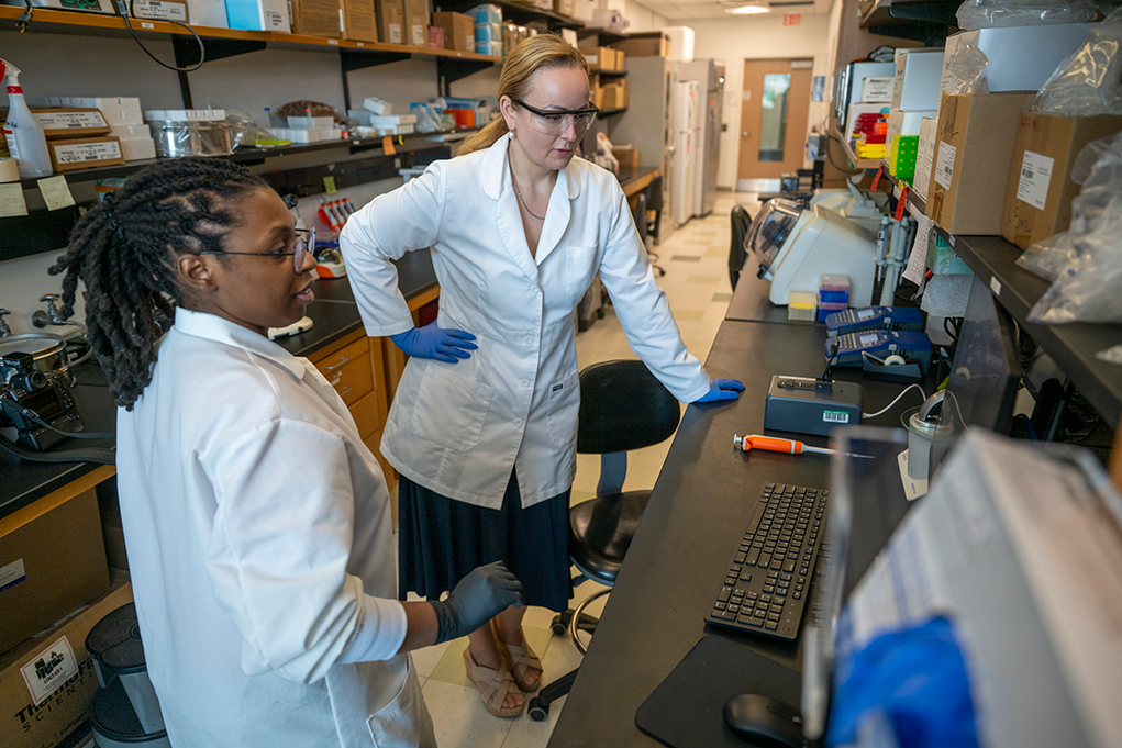 Two female scientists wearing lab coats, gloves, and safety glasses look at a computer in a laboratory.