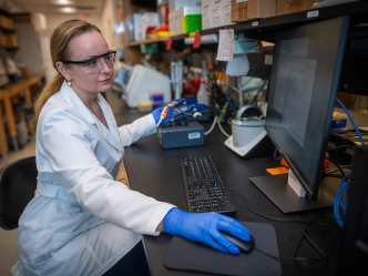 A woman wearing a lab coat and safety glasses sits at a computer in a laboratory and looks at the screen. She's wearing gloves and has one hand on the computer mouse and one hand holds a scientific instrument.
