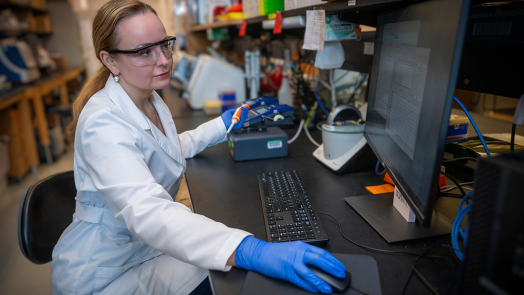 A woman wearing a lab coat and safety glasses sits at a computer in a laboratory and looks at the screen. She's wearing gloves and has one hand on the computer mouse and one hand holds a scientific instrument.