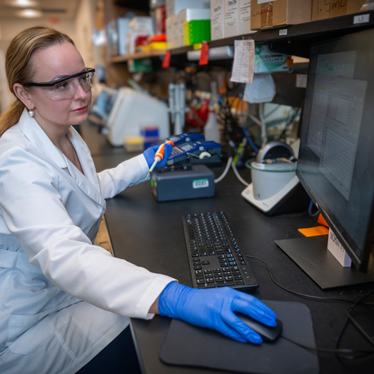 A woman wearing a lab coat and safety glasses sits at a computer in a laboratory and looks at the screen. She's wearing gloves and has one hand on the computer mouse and one hand holds a scientific instrument.