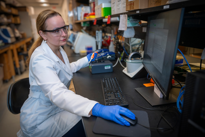 A woman wearing a lab coat and safety glasses sits at a computer in a laboratory and looks at the screen. She's wearing gloves and has one hand on the computer mouse and one hand holds a scientific instrument.