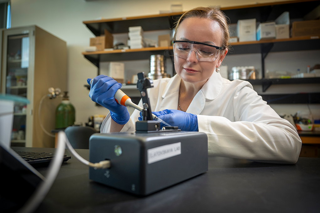 A female scientist wearing safety glasses, gloves, and a lab coat holds a scientific instrument and looks down at a table.