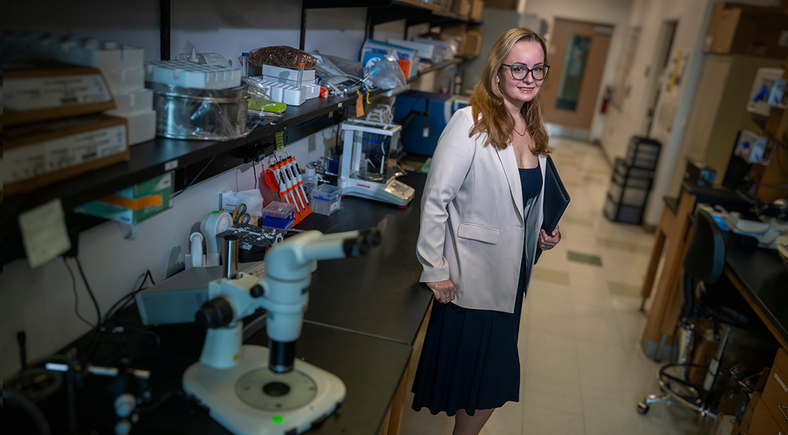 A woman wearing a lab coat stands with her back to a table in a science laboratory. She holds a folder and smiles at the camera.