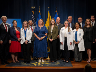 Men and women in business dress and white medical coats stand in front of flags.