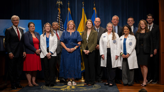 Men and women in business dress and white medical coats stand in front of flags.