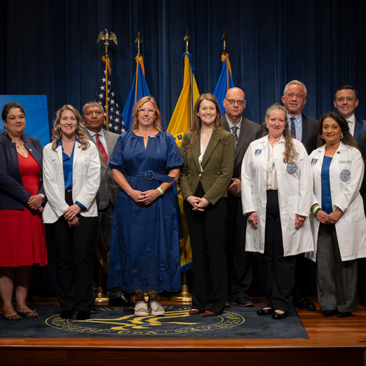 Men and women in business dress and white medical coats stand in front of flags.