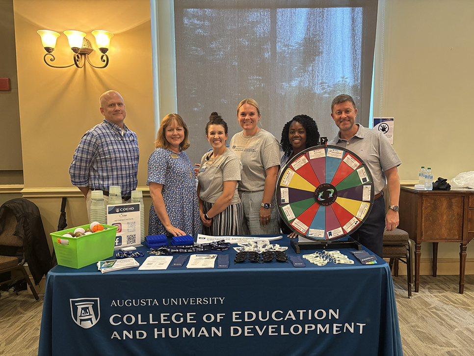 Men and women standing at a table with a game wheel in front of them. 