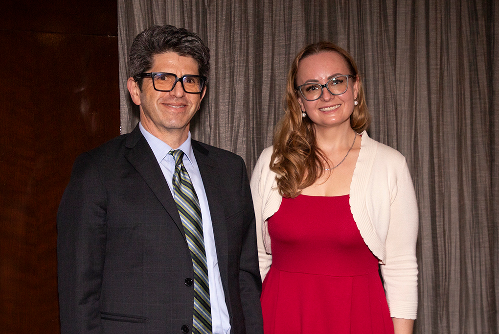A man wearing a suit and glasses and a woman wearing a dress and glasses smile at the camera.