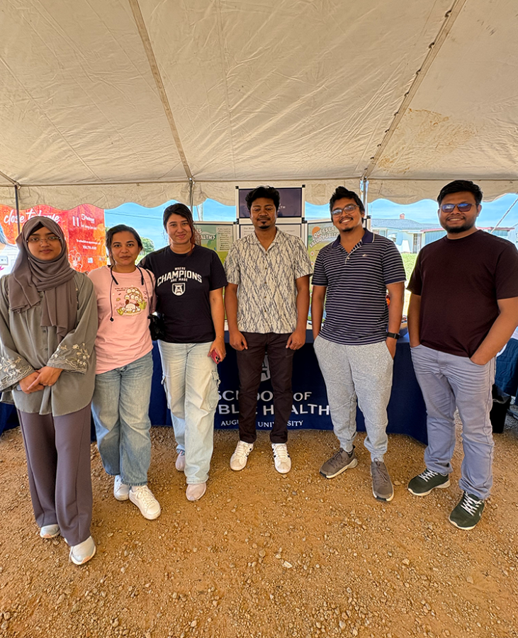 A group of PhD and master's students standing together under a tent at a health fair smiling at the camera.