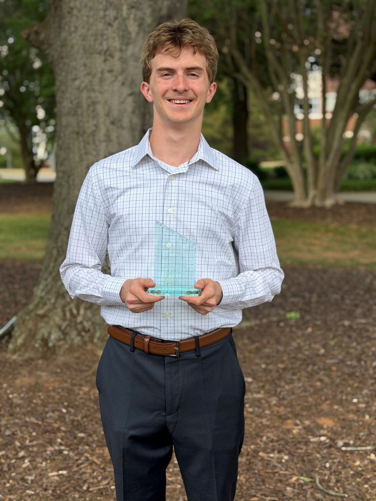 A college-aged boy standing outside while holding an award poses for a photo.