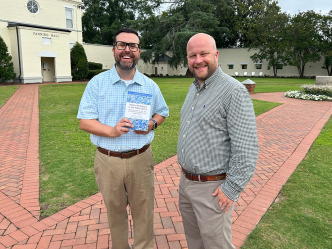 Two men standing on brick path, one holding a book.