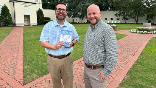 Two men standing on brick path, one holding a book.
