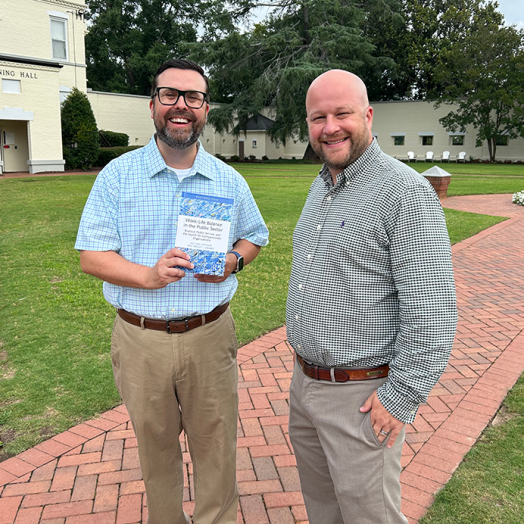 Two men standing on brick path, one holding a book.