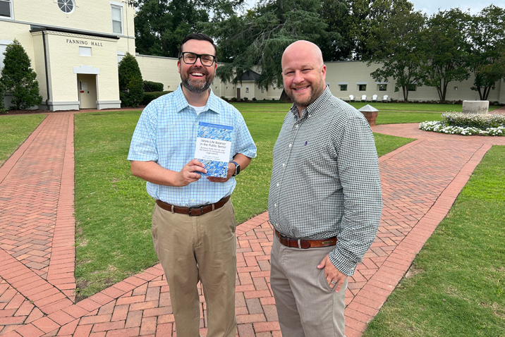 Two men standing on brick path, one holding a book.