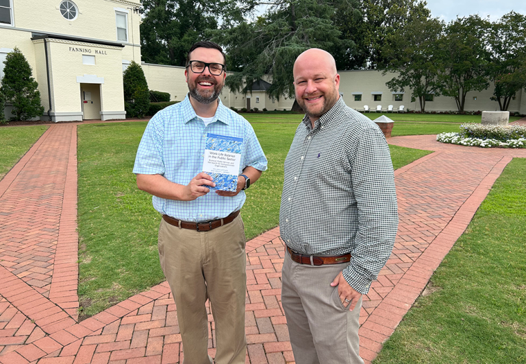 Two men standing on brick path, one holding a book.