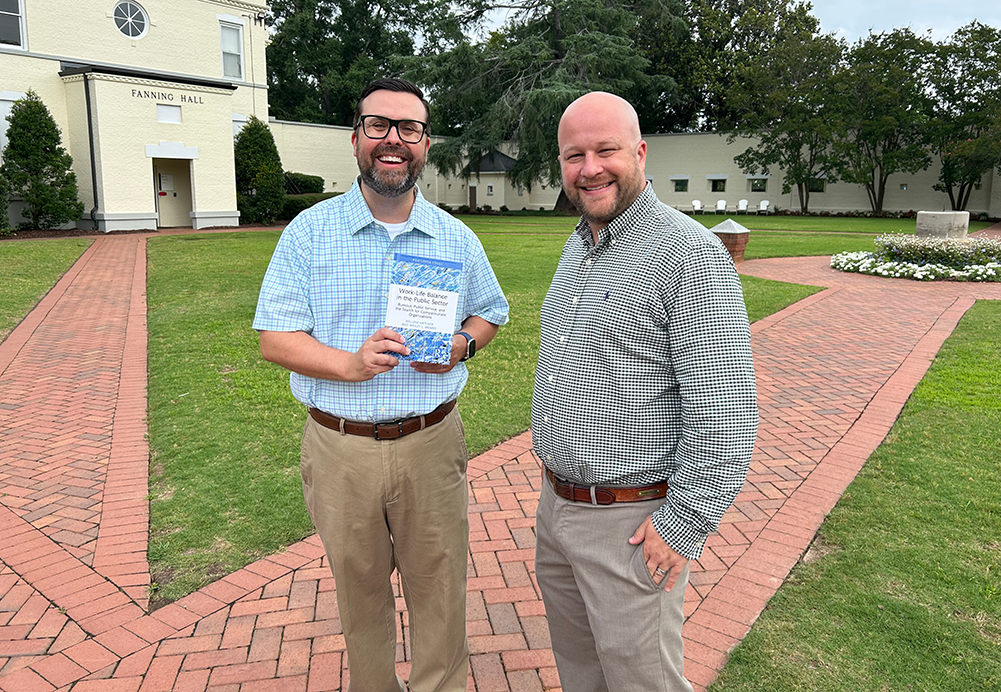 Two men standing on brick path, one holding a book.