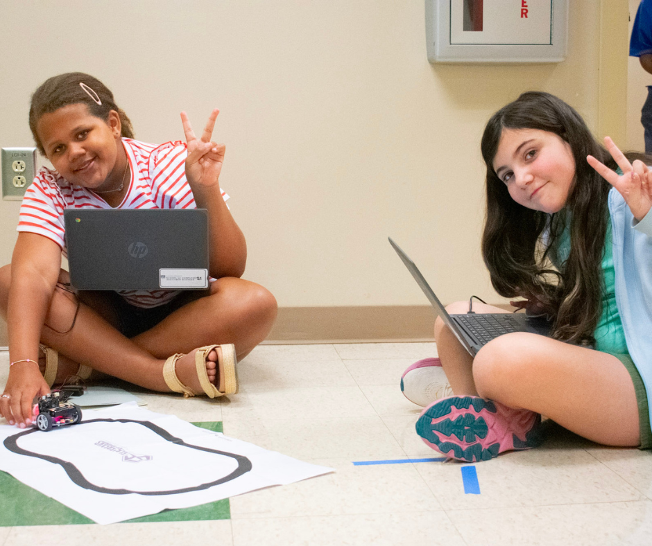 Students pose on the floor with their Micro:bit Cutebots, smiling and holding up peace signs.
