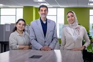 A male scientist and two female scientists stand at the end of a long table in a large, modern office space.