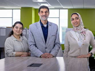 A male scientist and two female scientists stand at the end of a long table in a large, modern office space.