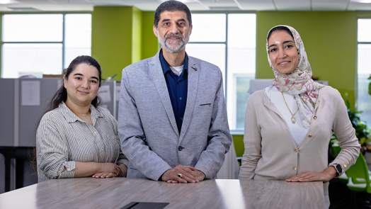 A male scientist and two female scientists stand at the end of a long table in a large, modern office space.