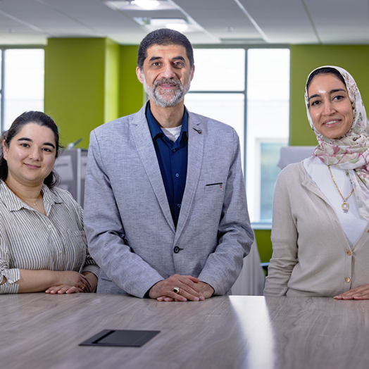 A male scientist and two female scientists stand at the end of a long table in a large, modern office space.